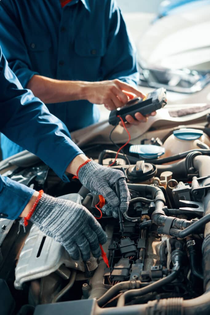 Deux mécaniciens testent un moteur de voiture dans un garage. L'un utilise des sondes sur les composants, l'autre manipule un multimètre pour le diagnostic.