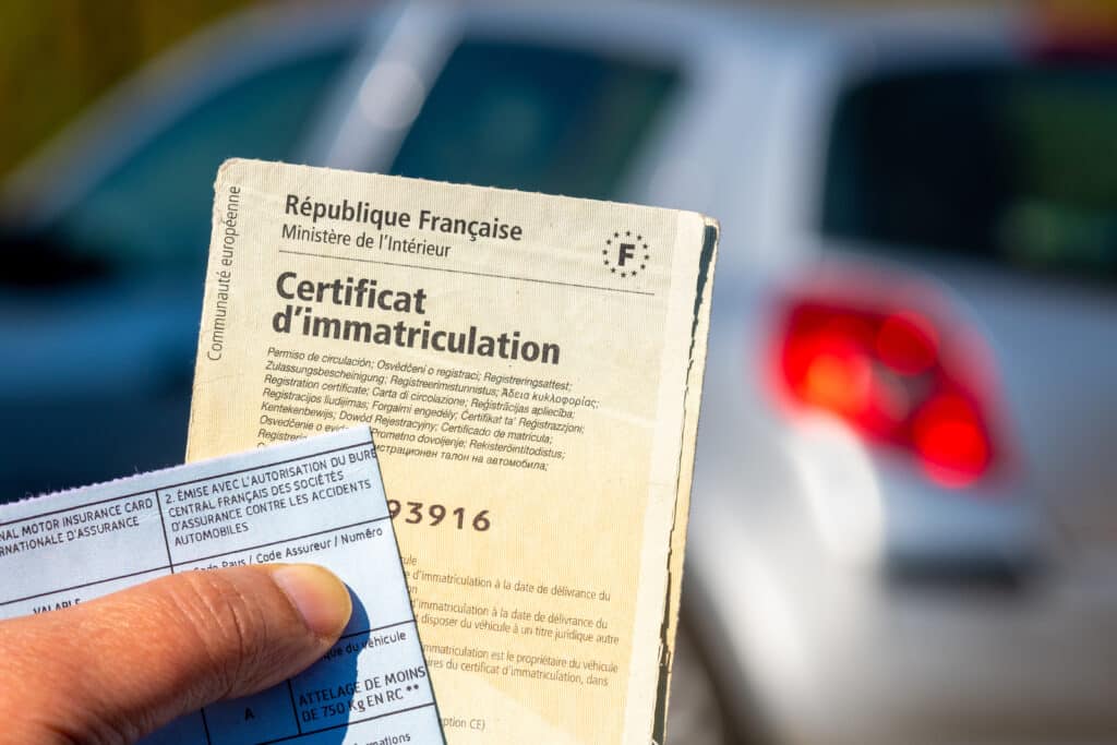 Main tenant le Certificat d'immatriculation français et une carte d'assurance auto. Texte "République Française". Voiture floue en fond.