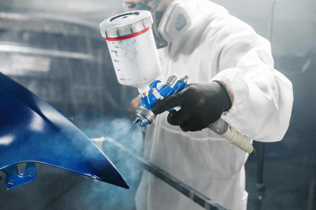 Opérateur en combinaison blanche et masque, pulvérisant de la peinture bleue sur une pièce de carrosserie automobile.