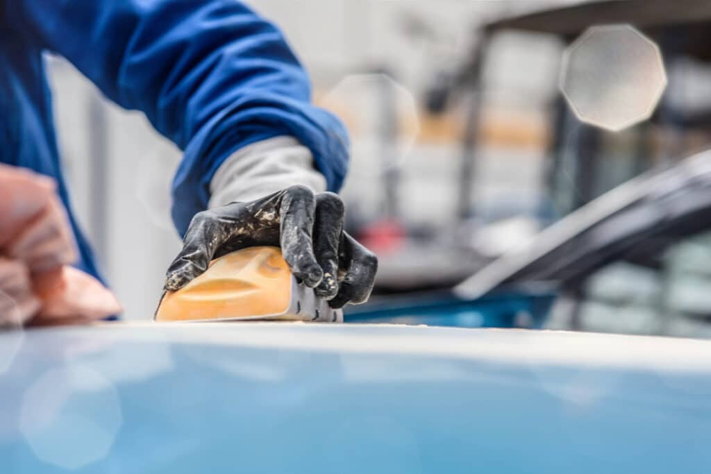 Ouvrier en gants noirs ponçant la carrosserie bleue d'une voiture avec un bloc de ponçage orange dans un atelier automobile.