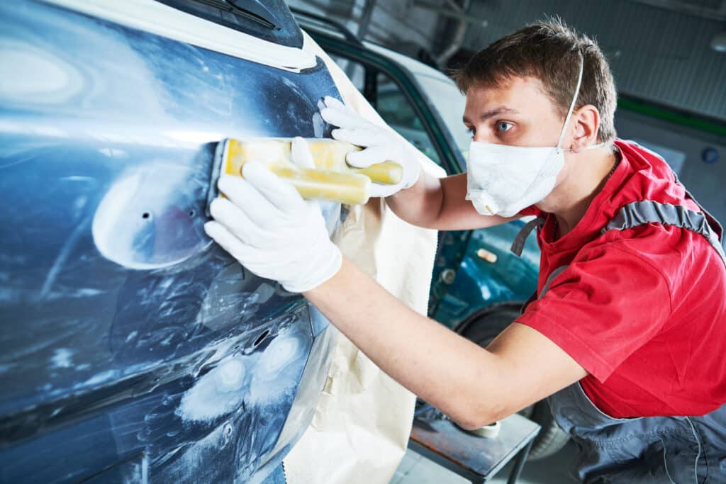 Carrossier masqué ponce la surface d'une voiture bleue avec un outil à main. Préparation minutieuse dans un atelier de réparation automobile.