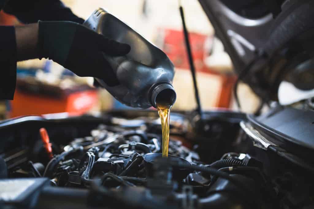 Main d'un mécanicien ganté versant de l'huile moteur dorée d'une bouteille grise dans le moteur ouvert d'une voiture, en garage.